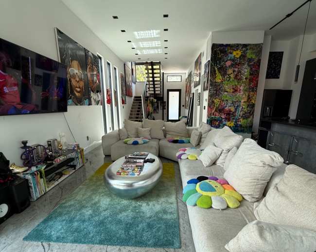 A modern living room features a series of large, light-colored sofas arranged around a central coffee table on a patterned rug, with walls adorned by colorful artwork and multiple televisions.