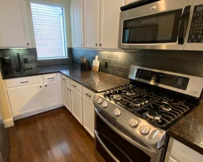A modern kitchen with stainless steel appliances, dark countertops, white cabinets, and a window letting in natural light.