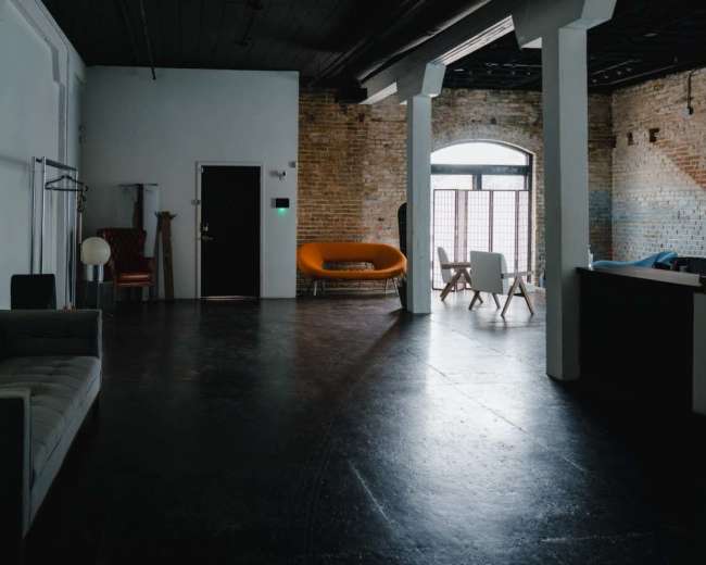 The image shows a spacious, minimalist room with exposed brick walls, featuring a gray sofa, an orange couch, and various furniture arranged in different areas.