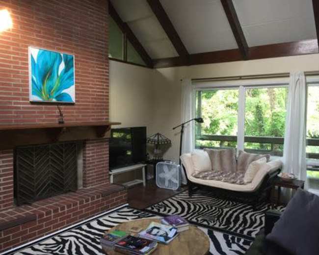 A living room with a brick fireplace, a large window overlooking greenery, a patterned rug, and a sectional sofa.