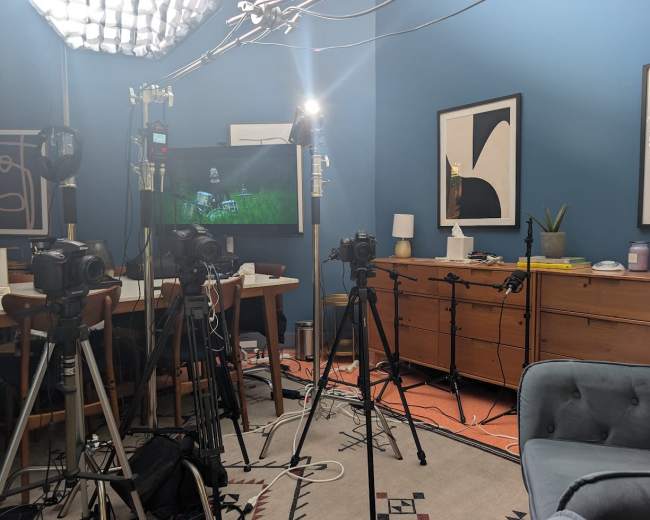 The image shows a room equipped for filming, featuring multiple cameras on tripods, lighting equipment, and a large screen displaying an image, with a mix of furniture and decor in the background.