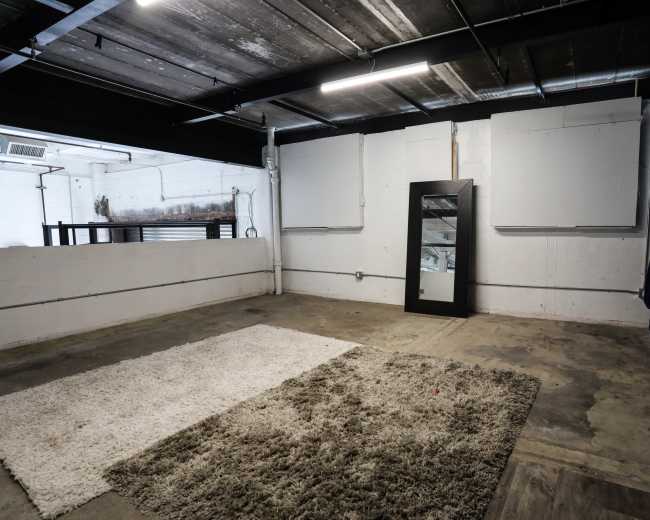 An empty room with a concrete floor, a large mirror, a dark curtain, and a plush rug in the center.