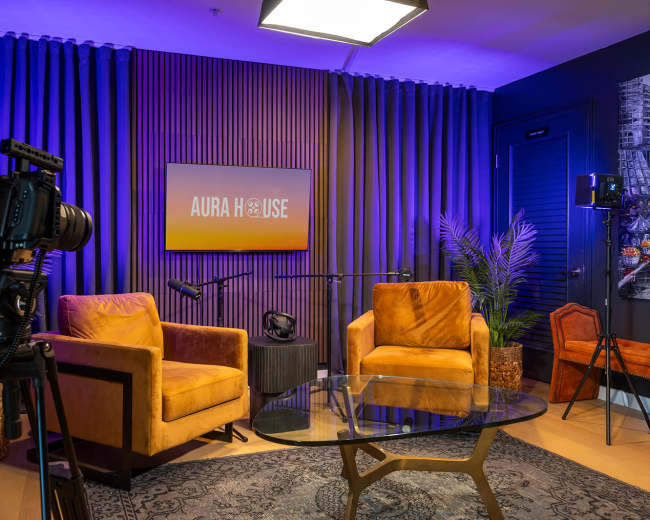 The image shows a modern studio setup with two orange chairs, a glass coffee table, and a large screen displaying "AURA HOUSE" against a backdrop of purple curtains and decorative plants.