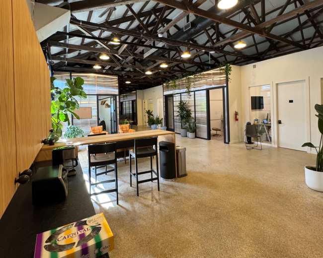 The image shows a spacious office interior with exposed wooden beams, a kitchen area with stools, and various plants scattered throughout the room.