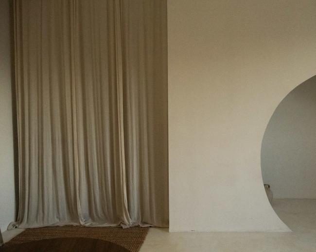 The image shows a minimalist interior space featuring a draped curtain on the left and a curved archway on the right against a plain wall.