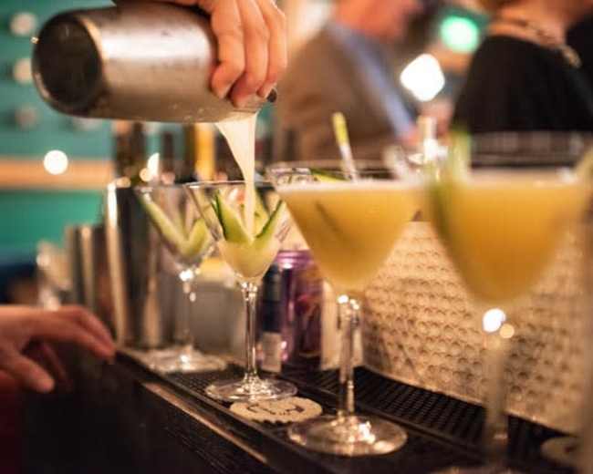 A bartender pours a yellow cocktail from a shaker into a martini glass garnished with cucumber at a bar.