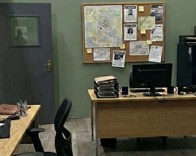 The image depicts a small office setup with two desks, a computer on each, a bulletin board with maps and notices, and a filing cabinet against the wall.