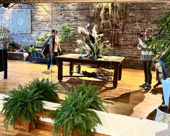 The image shows a spacious room with a mirrored wall, featuring a wooden table surrounded by plants and decorative elements, along with two people interacting in the space.