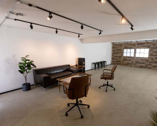 The image shows a minimalist interior space featuring a leather sofa, wooden table with two chairs, and a decorative plant, with a wall of wooden panels in the background.