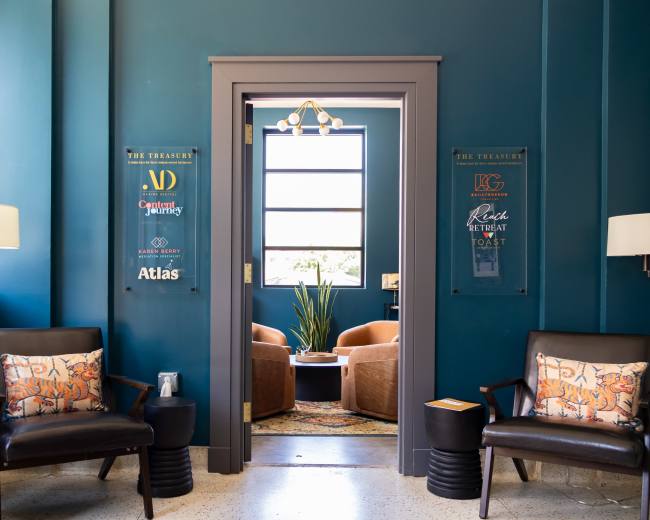 The image shows a cozy waiting area with two dark chairs, a potted plant, and a colorful rug, framed by a doorway leading to a brighter room with soft seating.