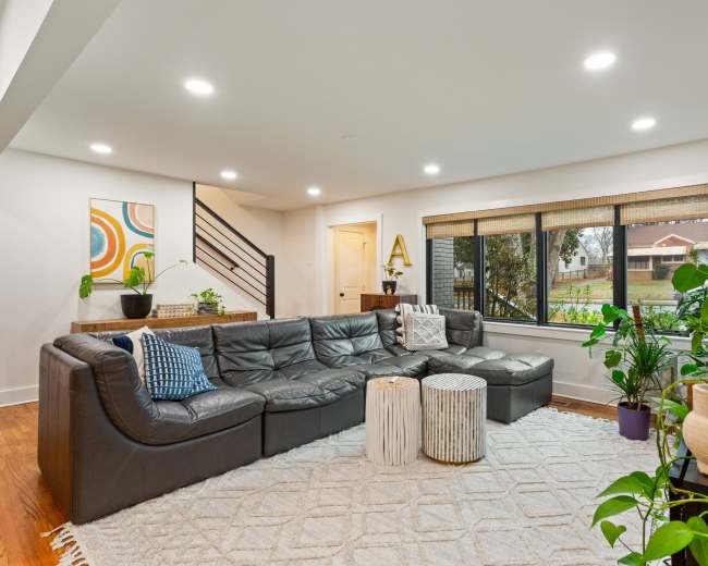 A modern living room features a large sectional sofa, a coffee table, decorative plants, and a window with a view of a garden.