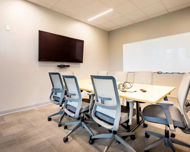 A modern conference room with a long table surrounded by six ergonomic chairs, a wall-mounted television, and a whiteboard.