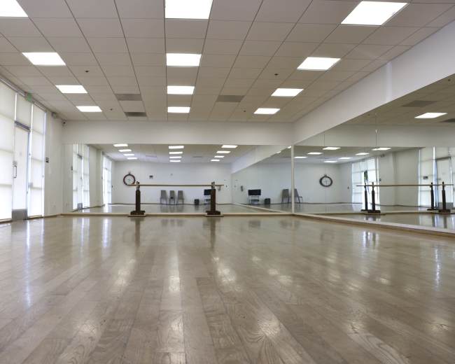 A bright, empty dance studio features wooden flooring, mirrored walls, and a ballet barre.