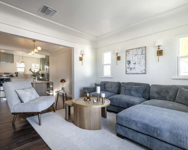 The image shows a modern living room featuring a gray sectional sofa, a round wooden coffee table, and an armchair, with an open view into a kitchen area in the background.