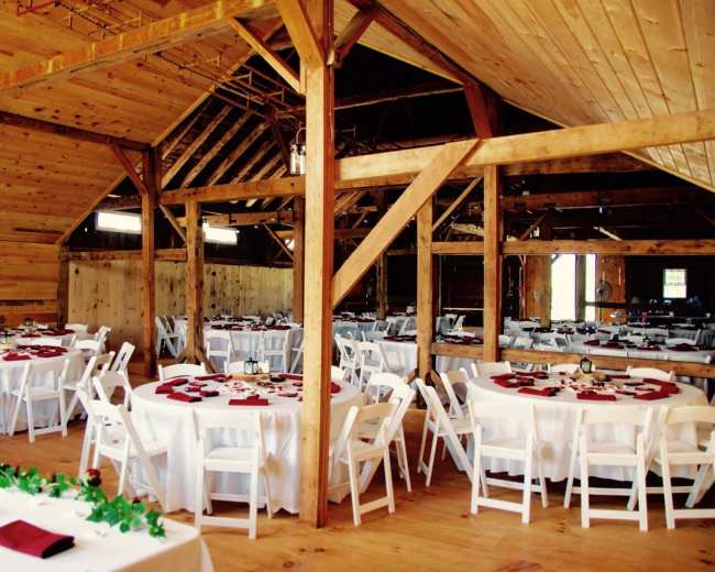 A spacious barn interior set up for an event, featuring several round tables with white tablecloths and red napkins.