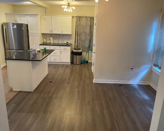 The image shows a kitchen and living area with wooden flooring, white cabinetry, and a stainless steel refrigerator.