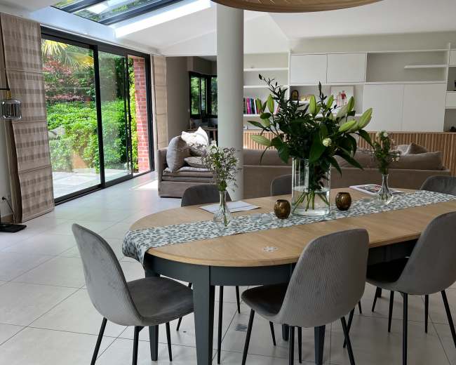 A modern dining area with a wooden table, grey chairs, and a large light fixture, set against a backdrop of large glass windows overlooking a garden.