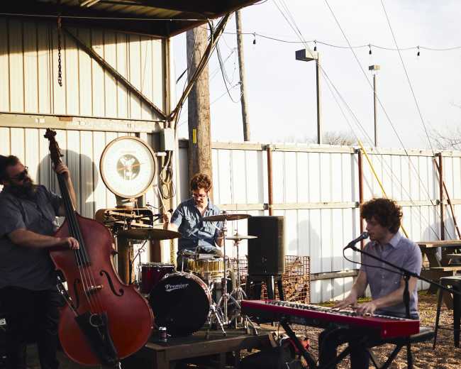 Three musicians perform with a double bass, drums, and a keyboard in an outdoor venue under a metal roof.