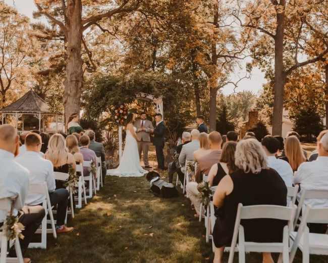A couple stands under an arch surrounded by trees while guests seated on white chairs observe their wedding ceremony.