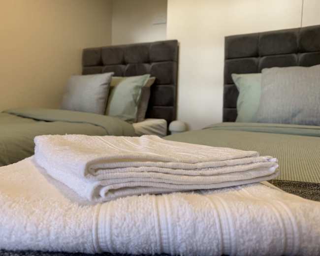 The image shows two neatly made beds with gray headboards and stacked white towels in the foreground.