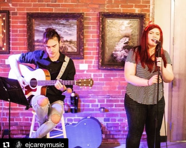 A man plays acoustic guitar while a woman sings into a microphone, with a brick wall and framed pictures in the background.