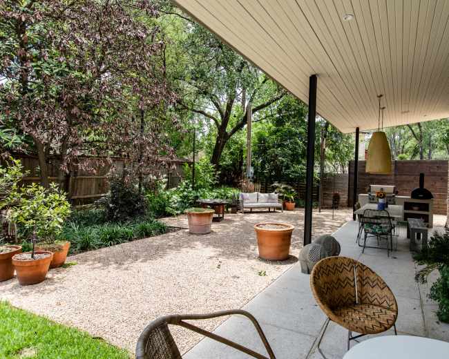 The image shows a landscaped outdoor patio area featuring a seating arrangement, potted plants, and a grill under a modern overhang.
