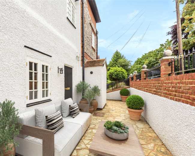 The image shows a small outdoor seating area beside a white house, featuring a sofa, potted plants, and a stone pathway.