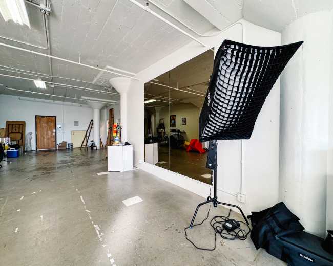 The image shows a spacious, unfinished studio with a large mirror, lighting equipment, and scattered furniture.