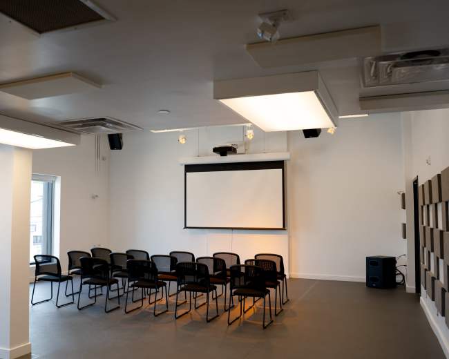 A meeting room features a row of black chairs arranged facing a large screen on the wall, with a projector mounted above.