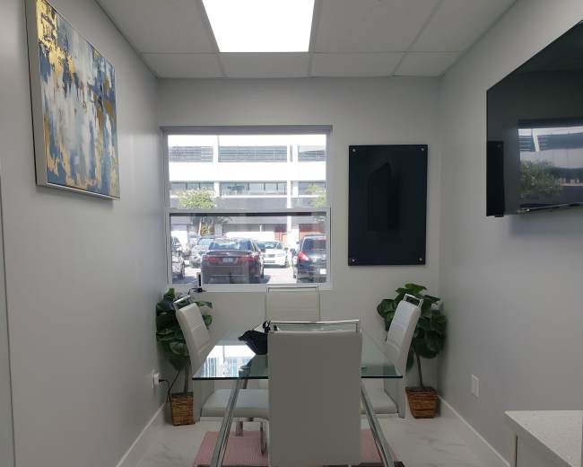 A small conference room with a glass table surrounded by four white chairs, and a window overlooking a parking lot.