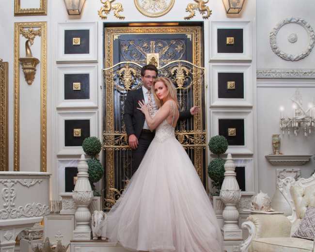 A couple poses in elegant formal wear in front of an ornate gold and black entrance, surrounded by lavish decor.
