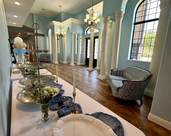 The image shows a decorated hallway with elegant columns, a table set with dishes and candles, and a seating area with a patterned chair.