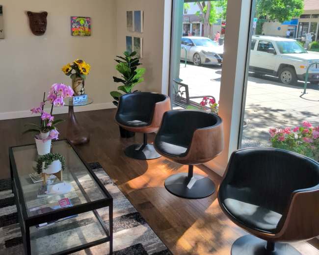 The image shows a well-lit interior space with three modern black chairs, a glass table, and decorative plants, overlooking a street with parked cars outside.
