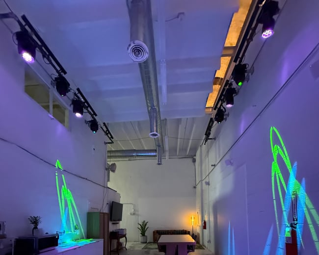 The image shows a spacious room with colorful lights projecting patterns on the floor and walls, featuring a large table in the center and a plant in the background.