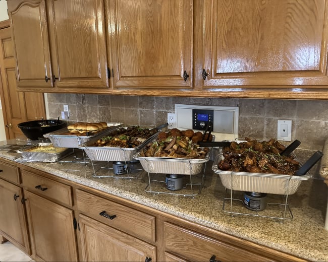 A kitchen counter with several large trays of assorted food, including meats, vegetables, and bread, arranged neatly in front of wooden cabinets.