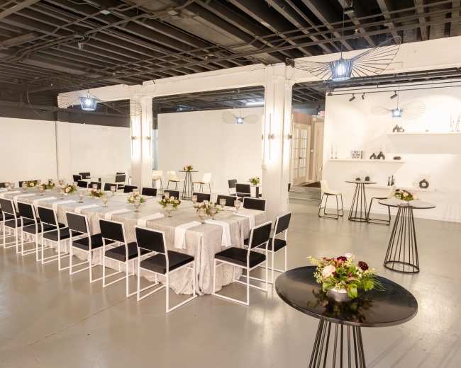 A spacious event room features a long table set for a dining arrangement with black chairs and floral centerpieces, surrounded by white walls and minimal decor.