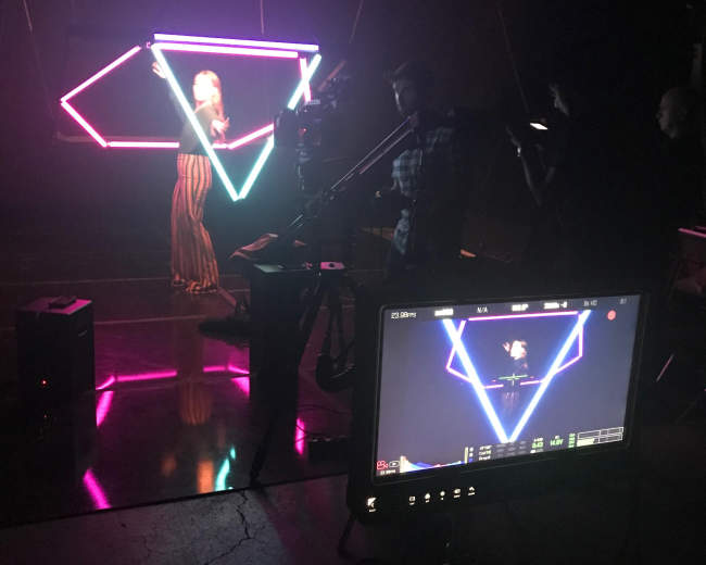 A person stands in front of a neon-lit geometric backdrop while a camera crew records from a monitor.
