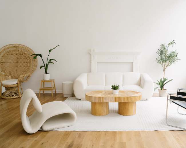 The image shows a modern living room featuring a white sofa, a round wooden coffee table, a pair of indoor plants, and a mix of minimalist furniture.