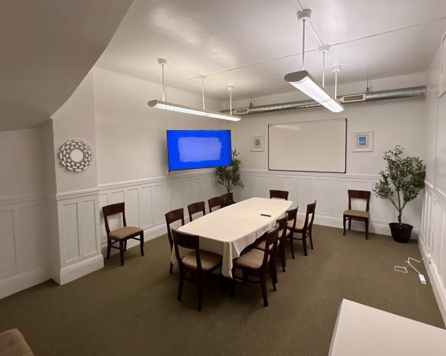 The image shows a meeting room with a rectangular table set for twelve chairs, a large screen on one wall, and a couple of potted plants in the corners.