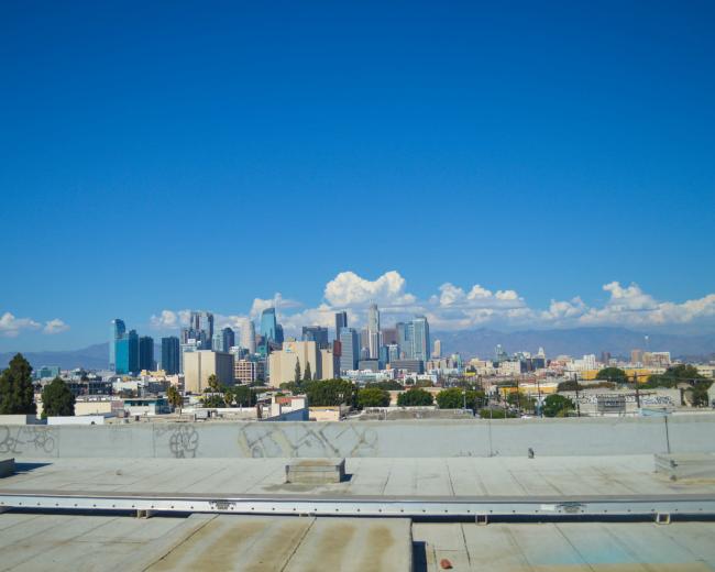 A clear view from a rooftop overlooks the skyline of downtown Los Angeles against a blue sky with scattered clouds.