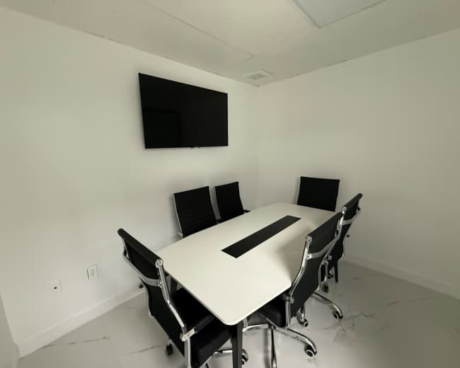 The image shows a modern conference room featuring a rectangular white table surrounded by six black chairs and a wall-mounted television.