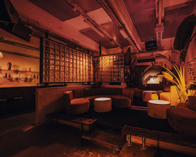 The image shows a dimly lit lounge area featuring circular seating, low tables, and a backdrop of decorative panels, all illuminated by warm orange light.