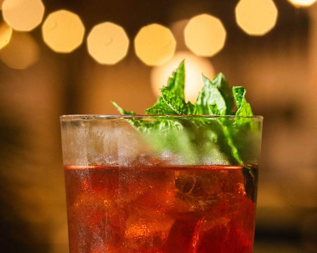 A glass of red cocktail with ice and a mint garnish is displayed against a blurred background of round lights.