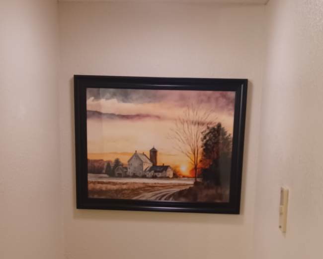 The image shows a bathroom with a white toilet and a framed landscape painting hanging on the wall above it.