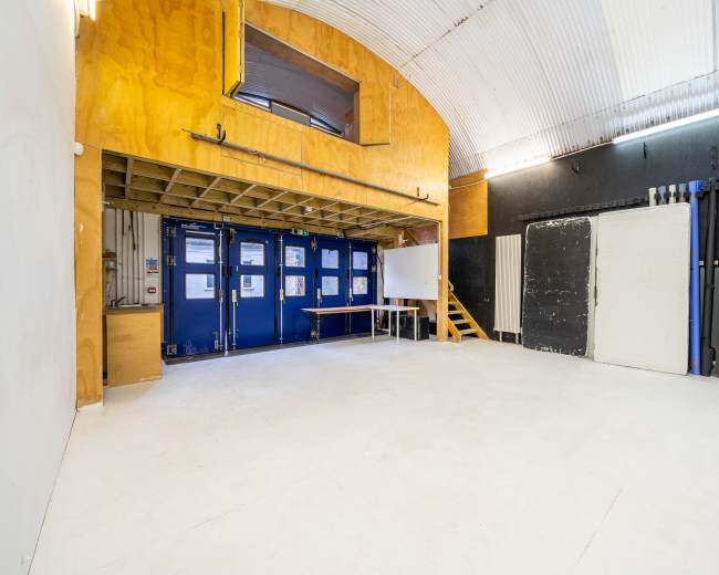 The image shows a spacious, empty studio room with wooden paneling and various colored materials stored against the wall.