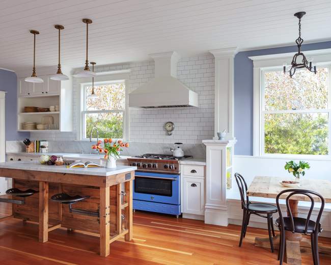 The image shows a modern kitchen featuring a wooden island, blue stove, white cabinetry, and large windows allowing natural light.