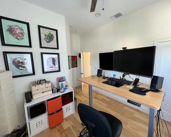 A modern home office features a standing desk with dual monitors, framed artwork on the wall, and organized storage units.