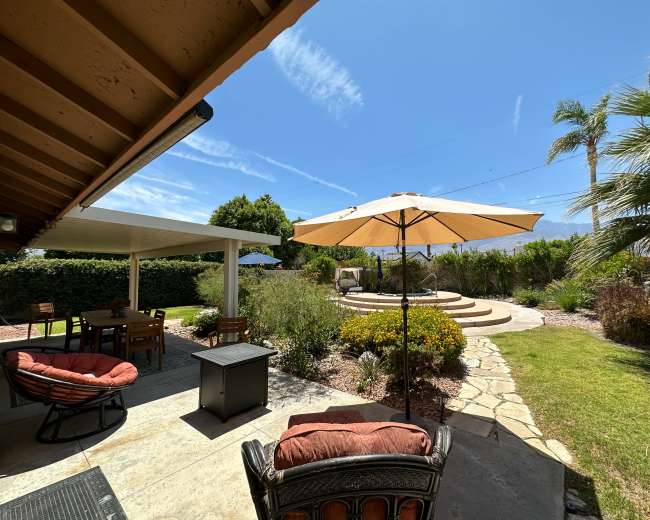 The image shows a landscaped backyard featuring a seating area with an umbrella, a stone pathway, and steps leading to a raised circular platform surrounded by greenery.