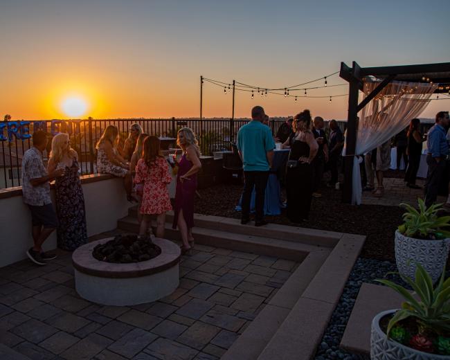 People are gathered on a rooftop celebrating at sunset, with decorations for a retirement event visible in the background.