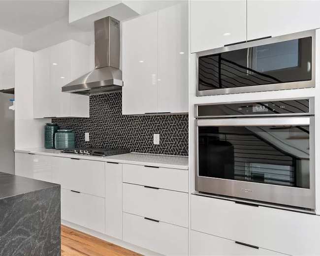 A modern kitchen featuring white cabinetry, a black hexagonal tile backsplash, stainless steel appliances, and a dark countertop.
